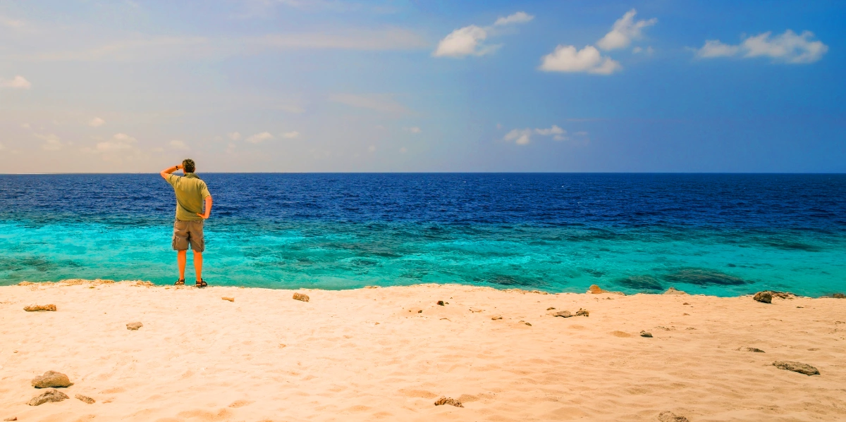 Man standing on sandy beach, hand shielding eyes, looking at turquoise ocean under a blue sky with scattered clouds