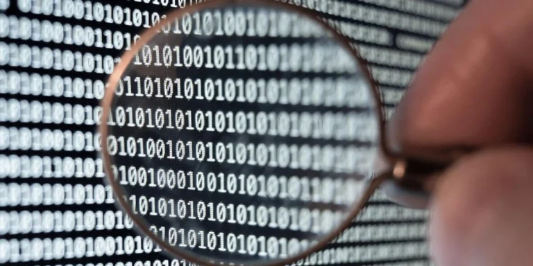 Magnifying glass held over binary code (0s and 1s) on a screen, symbolizing data inspection or analysis.