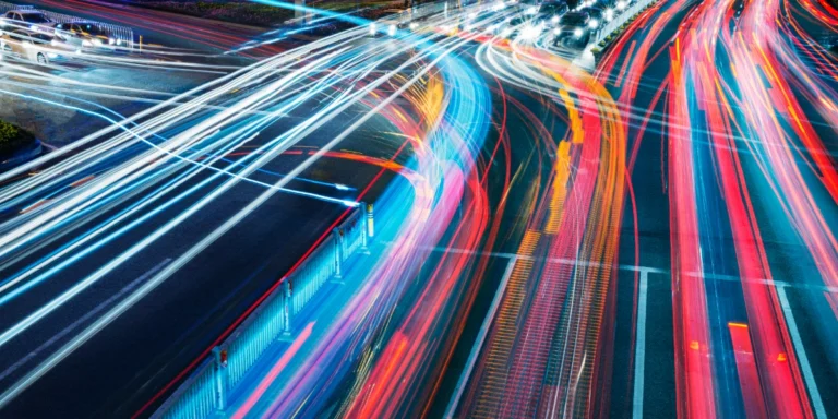 Long-exposure street scene showing colorful light trails from moving cars at a busy intersection at night, conveying speed and urban motion.