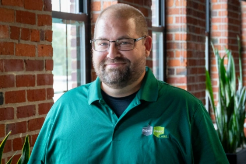 Smiling man with glasses in a green polo shirt indoors, brick walls and plants in the background.